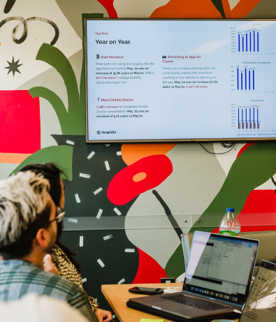 Two people, in a meeting, are viewing 'year on year' figures displayed on a screen.