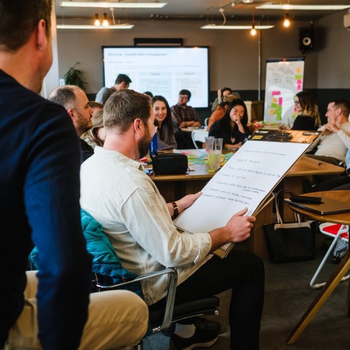 A room full of people. One person is holding large notepad and talking to the rest of the room with approximately 15 people other, all seemingly amused and enjoying the laid-back presentation