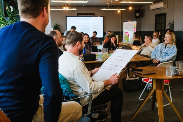 A room full of people. One person is holding large notepad and talking to the rest of the room with approximately 15 people other, all seemingly amused and enjoying the laid-back presentation