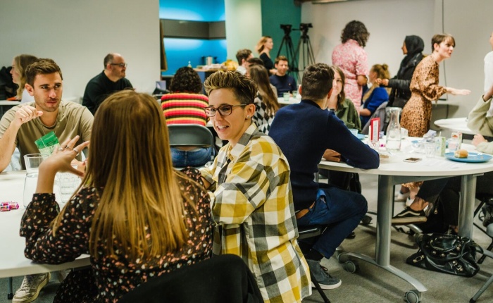 A room full of people, a team in a workroom which appears to be buzzing with conversation and idea sharing. Everyone seems to be fairly relaxed, in casual clothes but simultaneously engaged.