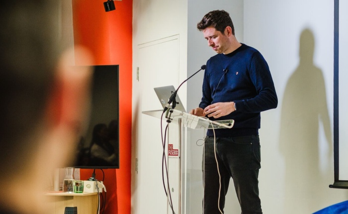 A man standing on a stage with a lecturn and on top is a laptop is resting on the table, and the person is additionally in front of a microphone, in a moment where he is pausing momentarily during his speech.