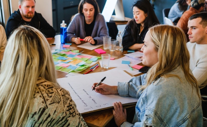 6 people around a table which is covered with neatly arranged postit notes, there is also a scribesperson who is writing some notes as the group ponder