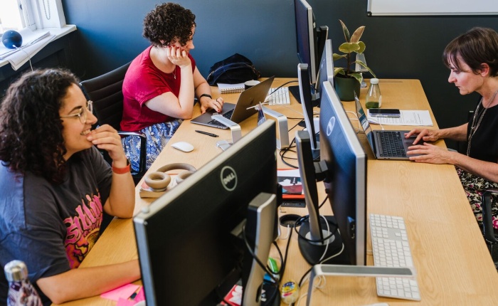 3 women sitting at a desk with laptops and monitors in front of them, in fairly casual clothes, looking intently at their screens as they seem to be working