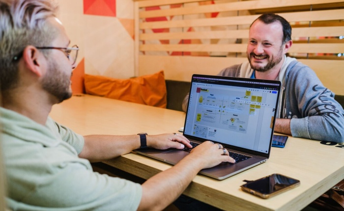 2 men sitting across from each other, one is smiling at the other whilst the other has his hands on a laptop, seemingly having an informal meeting