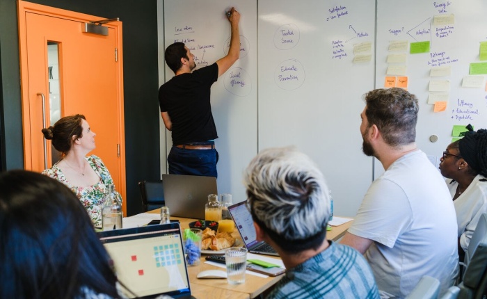 6 people sitting down at a large table with laptops but at the focal point of the image is 1 person standing and writing on a whiteboard whilst everyone watches