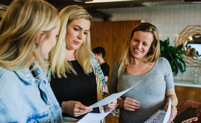 3 blonde women looking quite positive, staring at the piece of paper of the middle woman who is talking