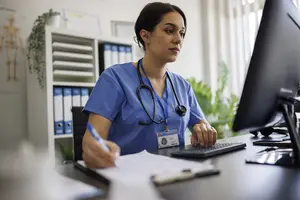 Professional female nurse or doctor working in a medical office, wearing blue scrubs and a stethoscope. She is typing on a computer keyboard and writing notes on a clipboard, concentrating on patient information or telemedicine documentation. The office background includes medical charts and organized files, conveying professionalism, care, and focus in a healthcare environment.