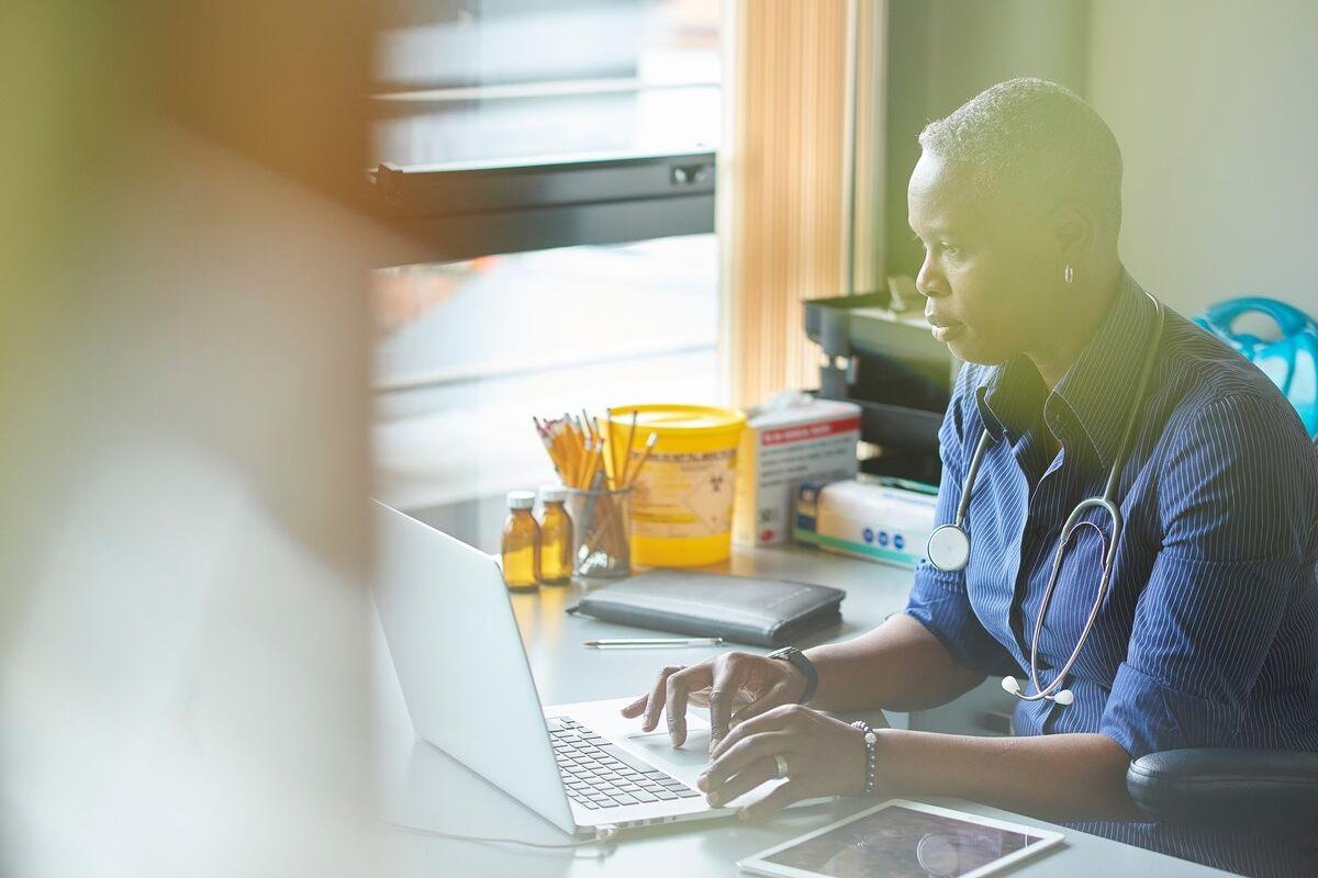 A medical professional with stethoscope round their neck sits at a desk and types on a laptop