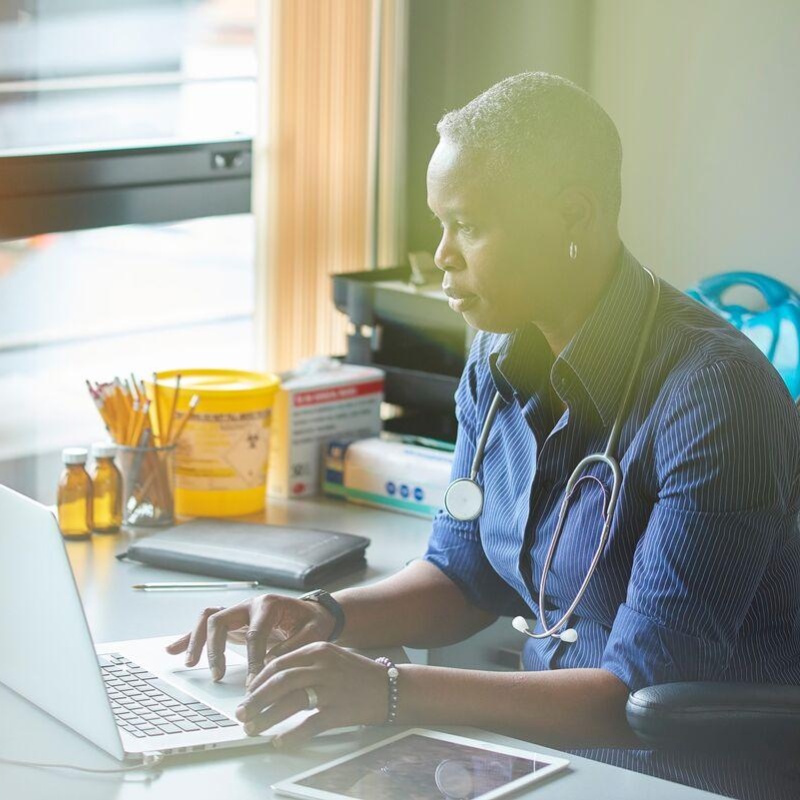 A medical professional with stethoscope round their neck sits at a desk and types on a laptop