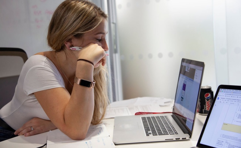 A person leaning on a desk to get closer to their laptop with a pen in their hand and a notebook on the desk