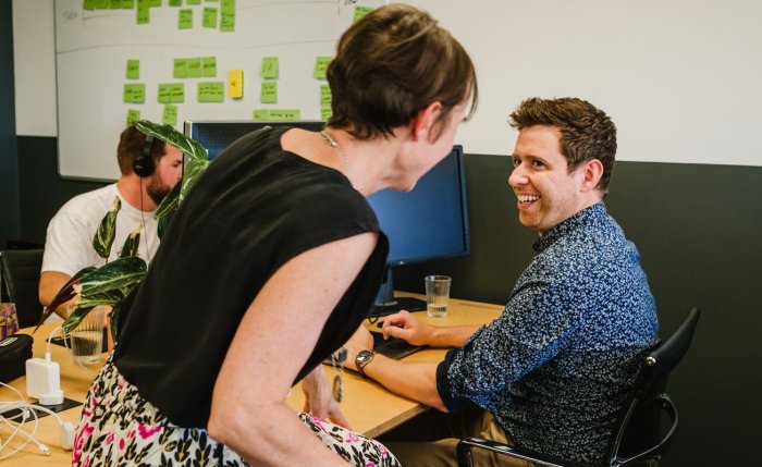 3 people in the back of an office, one standing up smiling, another sitting down smiling at the other and the third in the corner with headphones, discretely in the corner