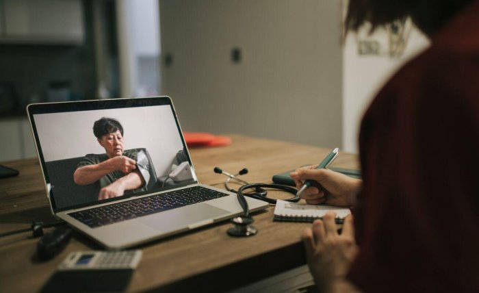 A slightly dark image with the focal point as a laptop with a patient who is taking their own blood pressure, unwrapping the apparatus around their arm and a doctor is in the forefront taking notes