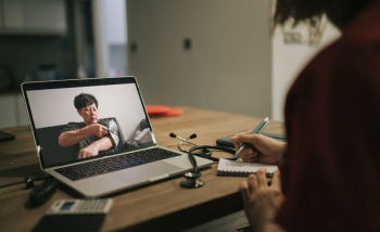 A slightly dark image with the focal point as a laptop with a patient who is taking their own blood pressure, unwrapping the apparatus around their arm and a doctor is in the forefront taking notes