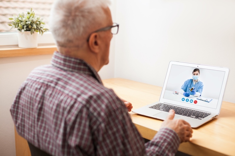 A doctor on a laptop screen, wearing blue scrubs and an older-looking patient talking to the doctor via video call whilst sitting down