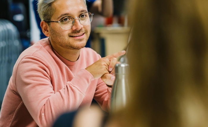 A person lightly smiling, wearing a pink jumper and glasses, looking at another person who is only indicated by a closeup of the back of their head.